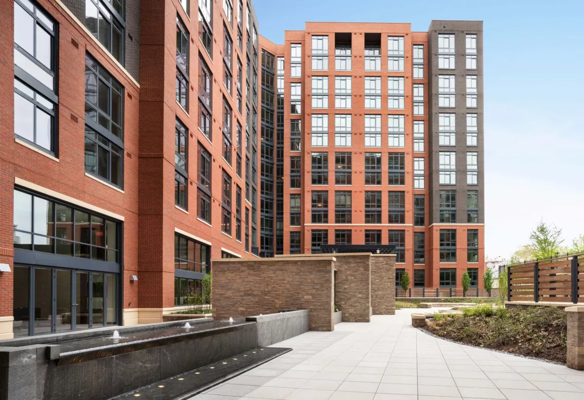 Residential courtyard walkway with concrete pavers, planters, and brick building walls.